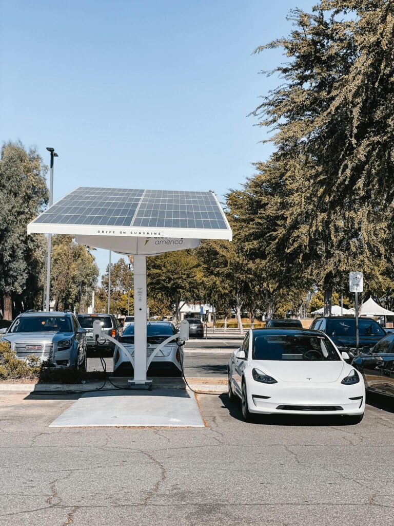 A white electric car charging under a solar panel in an urban setting, showcasing renewable energy use.